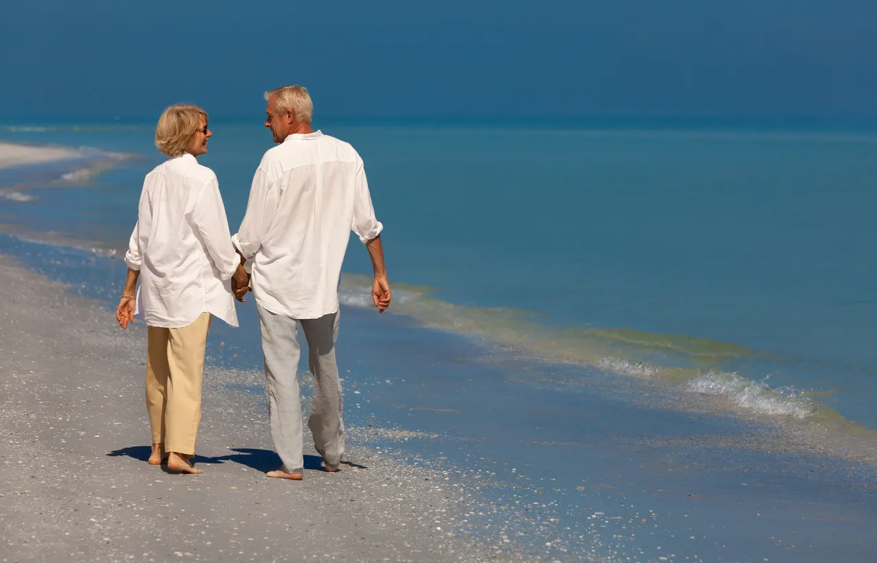 Happy senior man and woman couple walking smiling on vacation on a deserted tropical beach with bright clear blue sky and calm sea