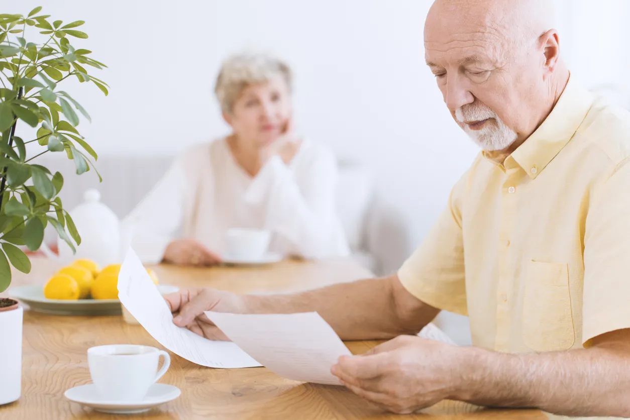 A worried senior sitting at a table and checking out papers and his wife looking at him